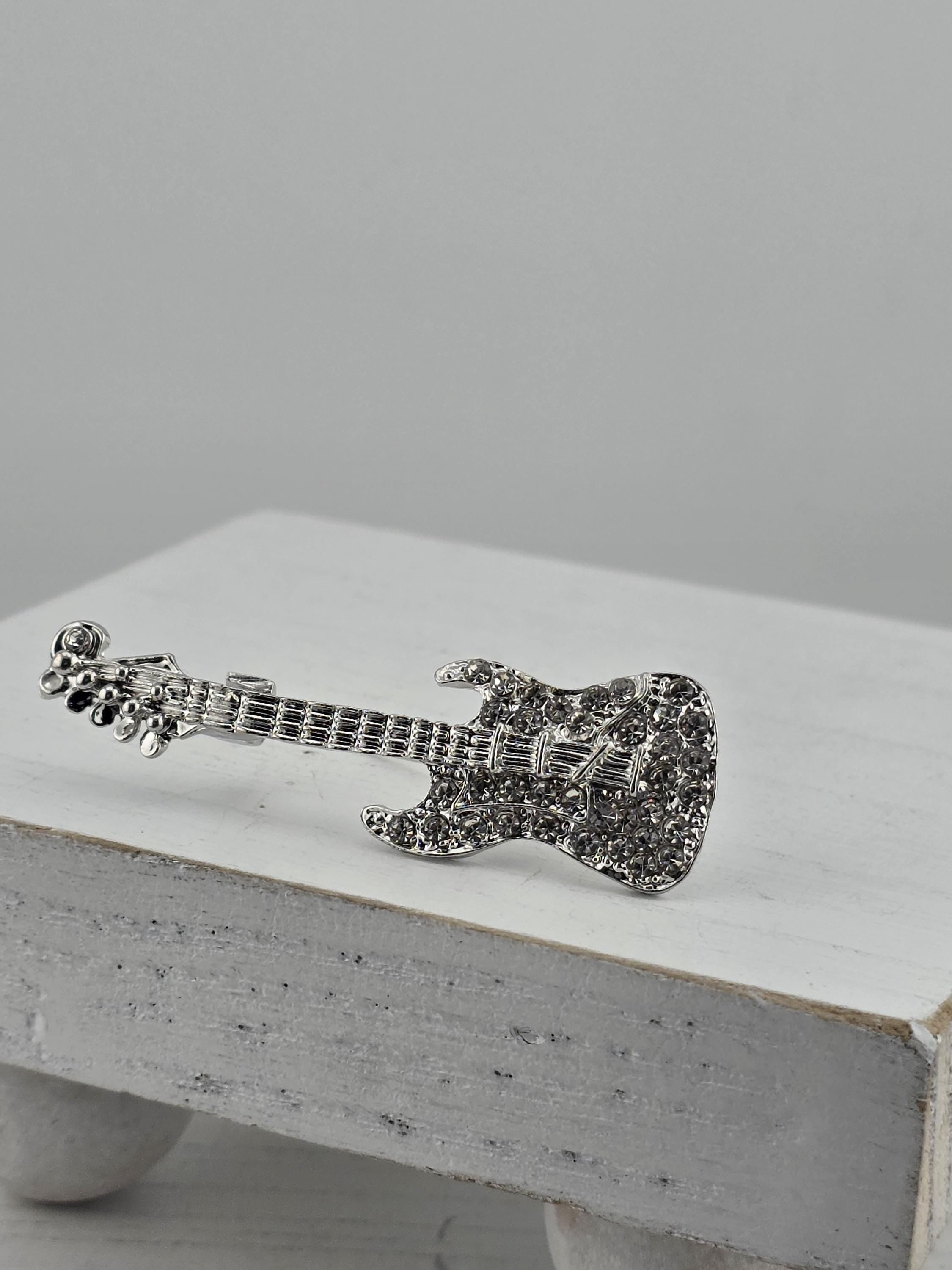 A silver-colored guitar brooch is displayed on a white surface.