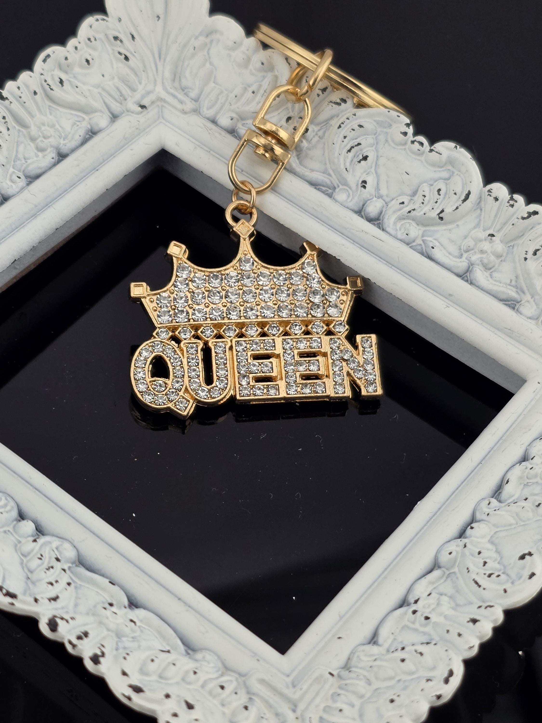 A gold-colored pendant with the word "queen" engraved on it, hanging from a white ornate frame.