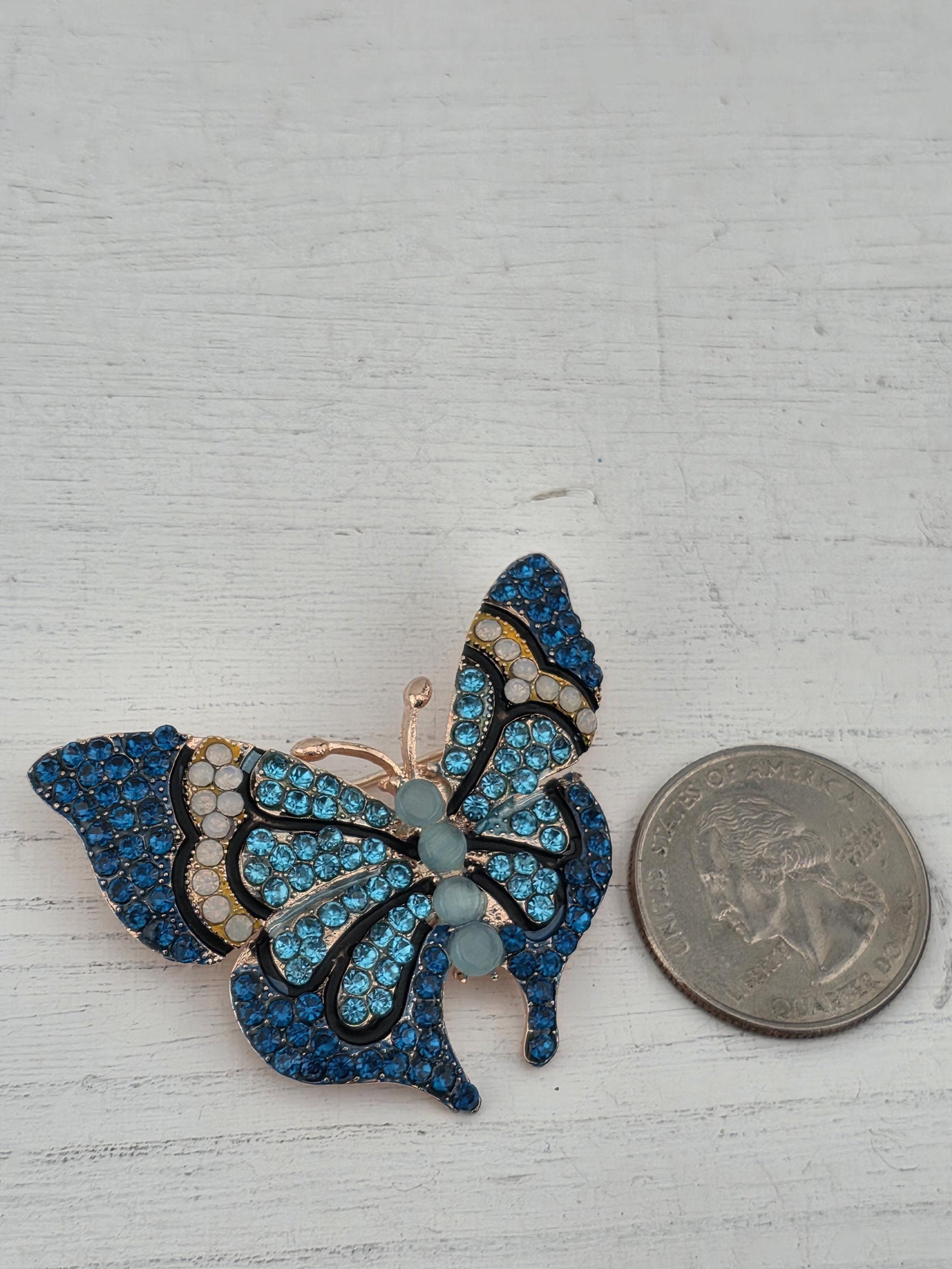A decorative butterfly brooch with blue and gold gemstones is placed next to a United States quarter on a white wooden surface.