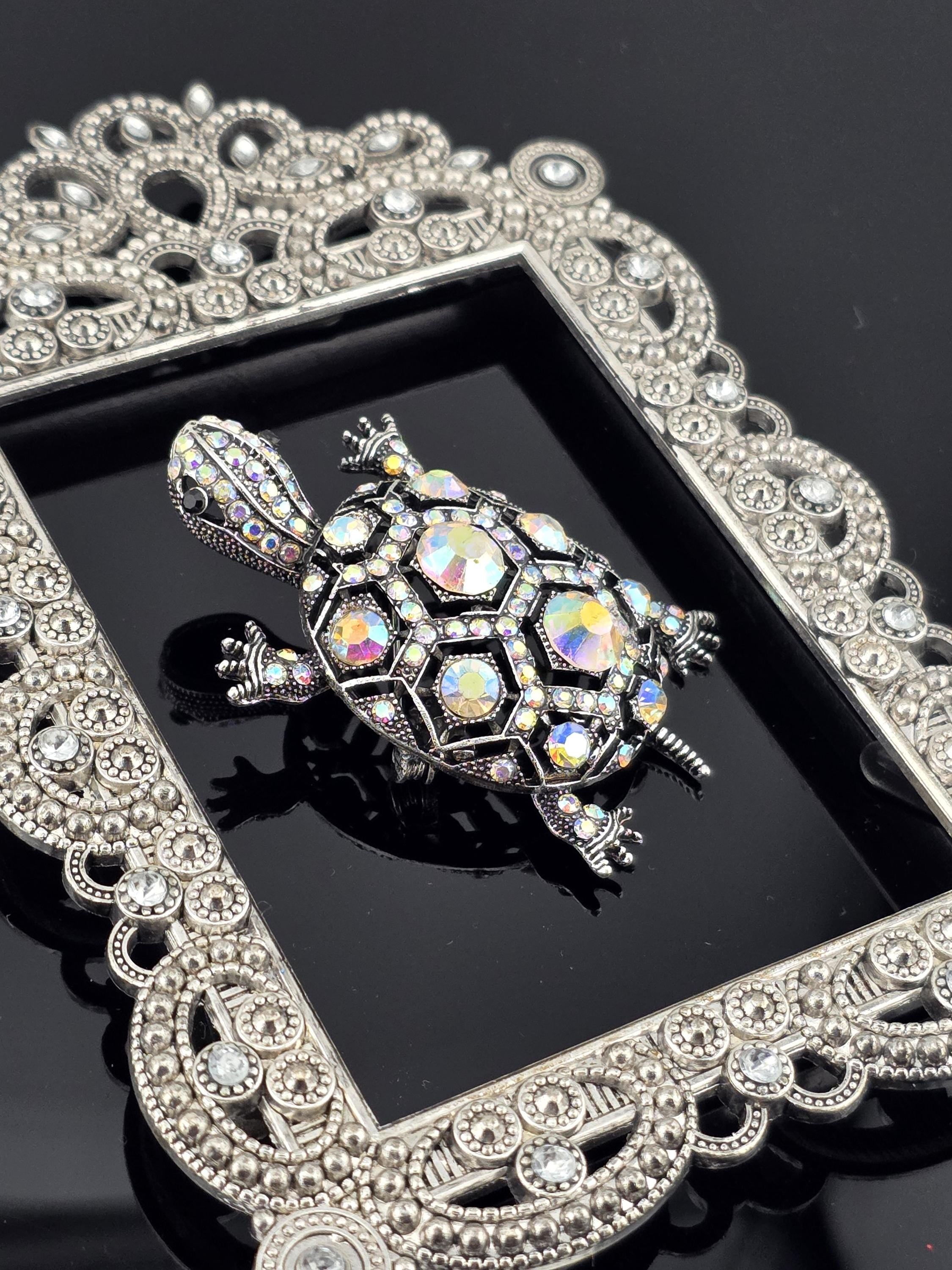 A decorative silver frame with a crystal turtle brooch is displayed against a dark background.