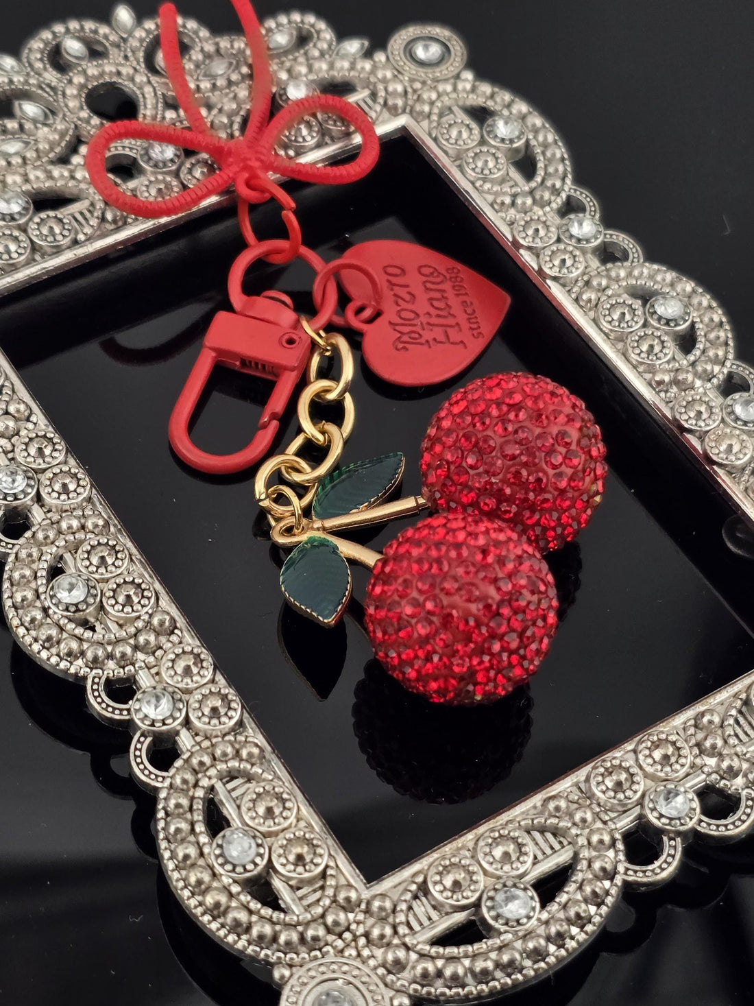 A decorative keychain with a red heart-shaped charm and two red crystal balls, placed on a black surface with a silver frame.