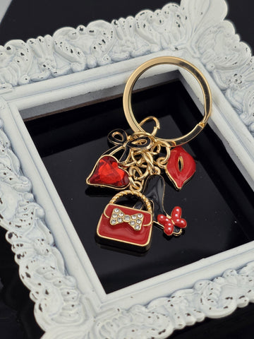 A decorative keychain with a gold frame and various colorful charms, including a red heart, a red flower, and a red bow, is displayed in a white ornate frame.