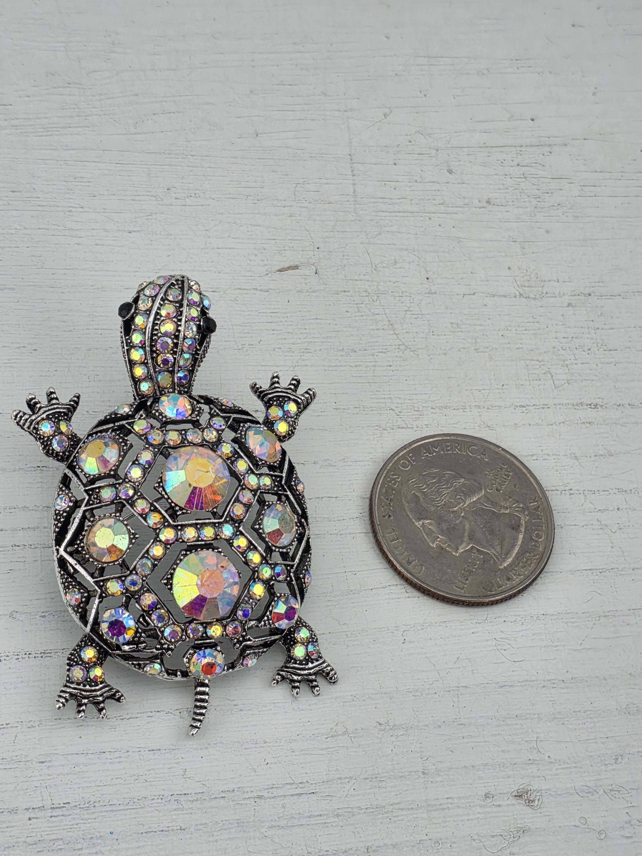 A silver-colored coin is placed next to a decorative turtle-shaped brooch adorned with colorful gemstones.
