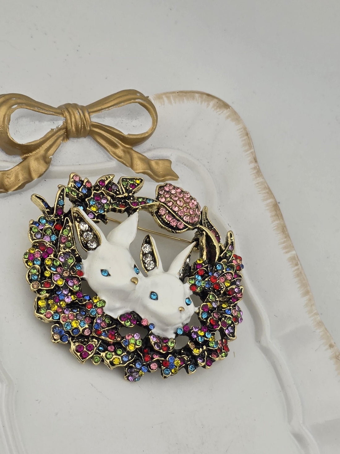 A decorative brooch with two white bunny heads, surrounded by a colorful floral wreath and a gold bow.