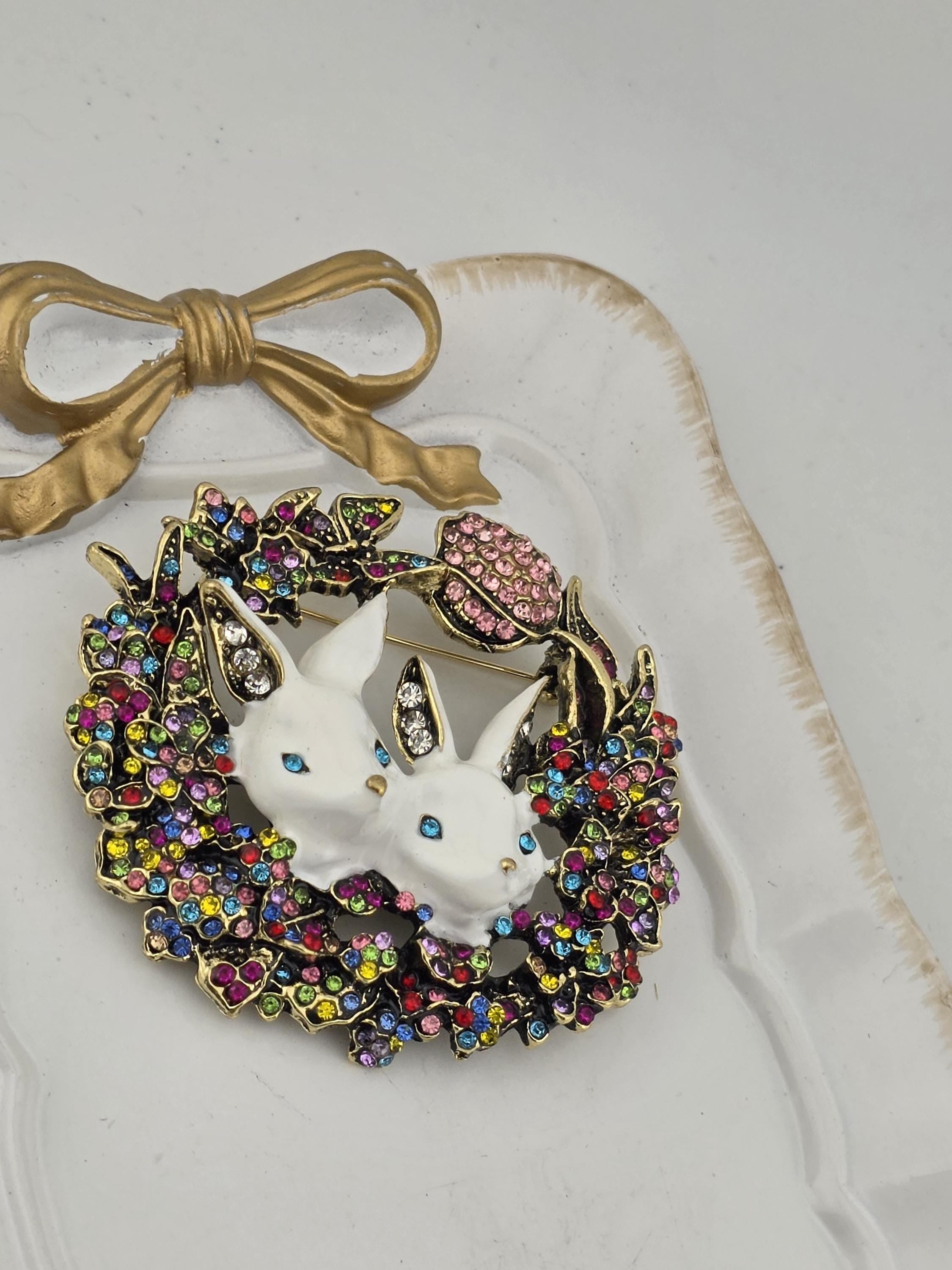A decorative brooch with two white bunny heads, surrounded by a colorful floral wreath and a gold bow.