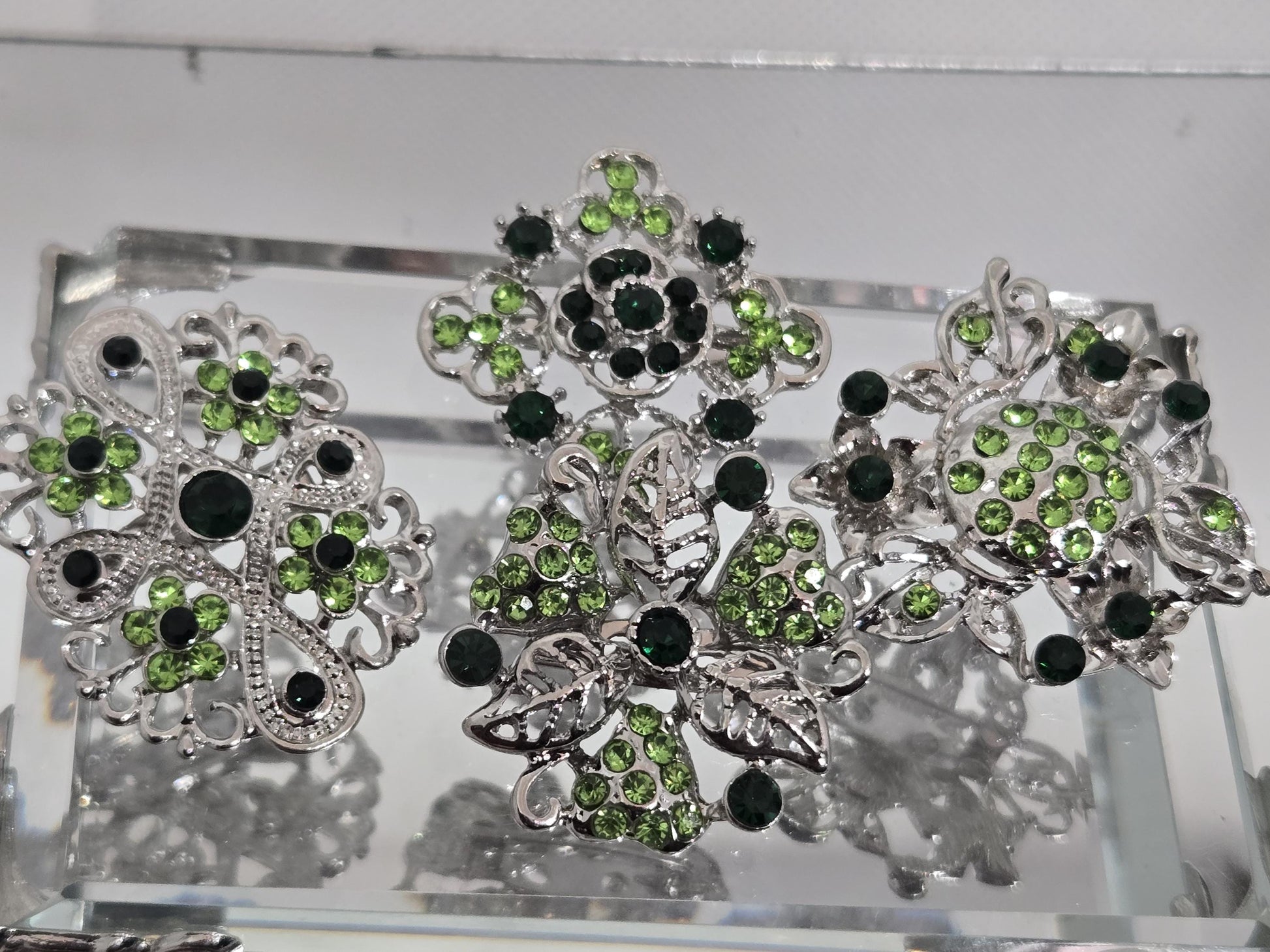 three ornate silver brooches adorned with green gemstones, including green diamonds and leaves, set in a decorative metal frame.