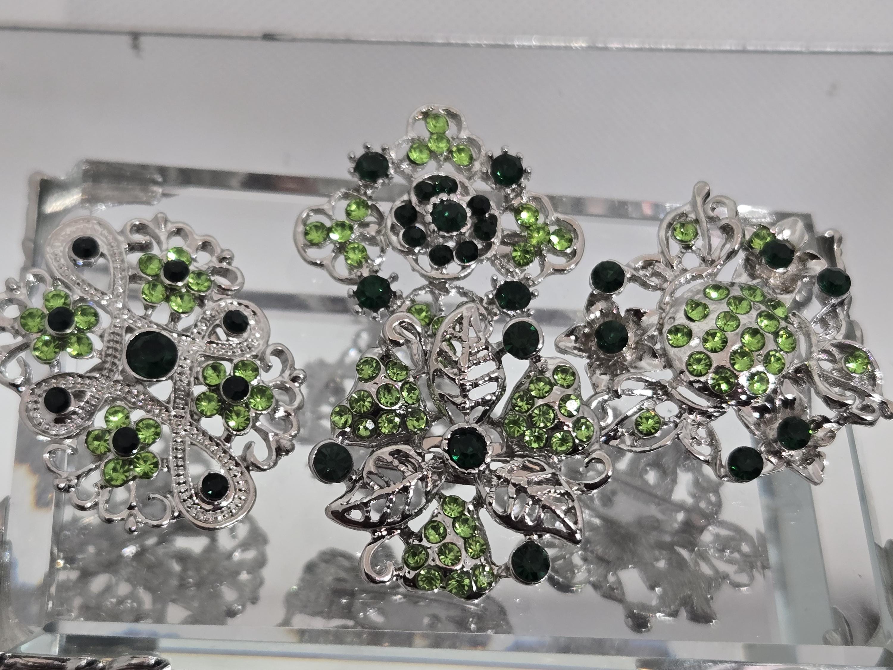 three ornate silver brooches adorned with green gemstones, including green diamonds and leaves, set in a decorative metal frame.