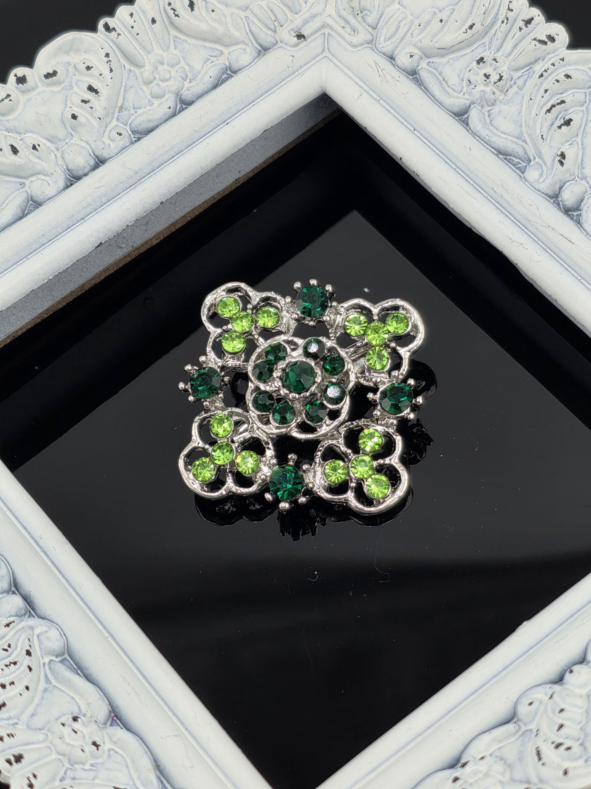 A decorative brooch with green and silver stones is displayed in a white ornate frame against a black background.