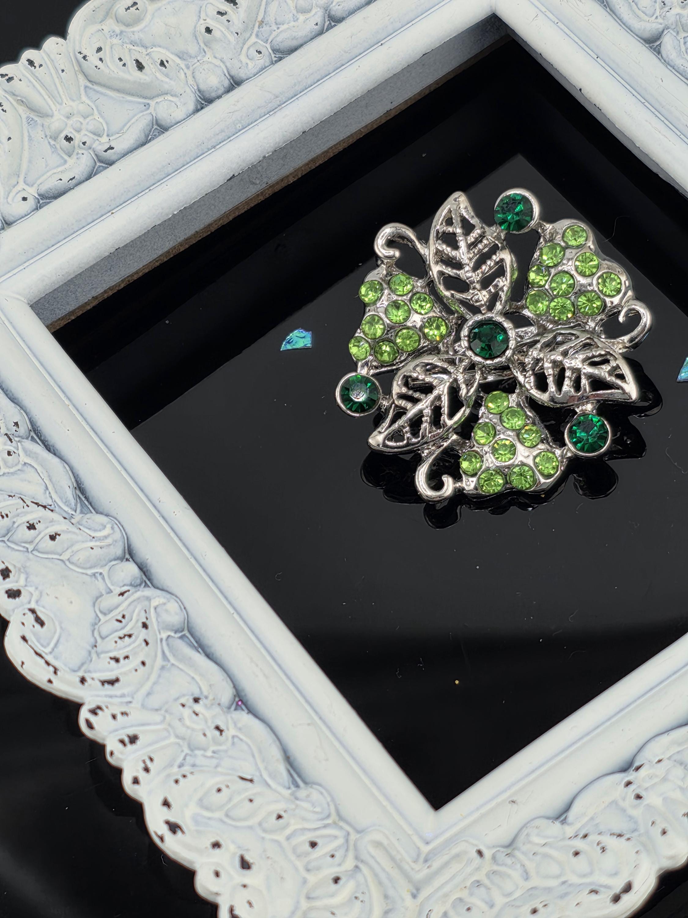 A decorative piece of jewelry with green and silver elements, displayed within a white ornate frame.