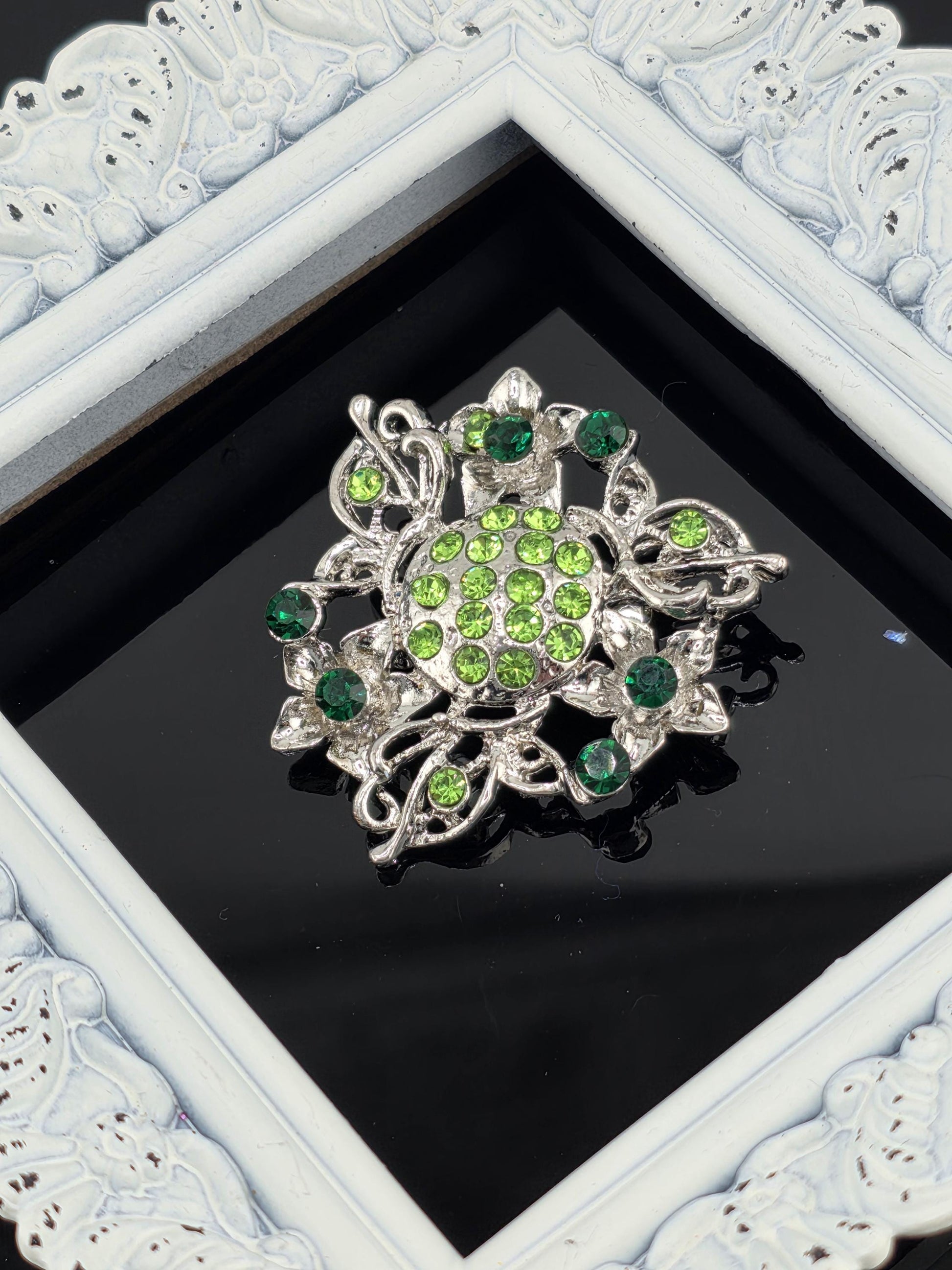 A decorative brooch with green gemstones and a silver frame is displayed against a black background.