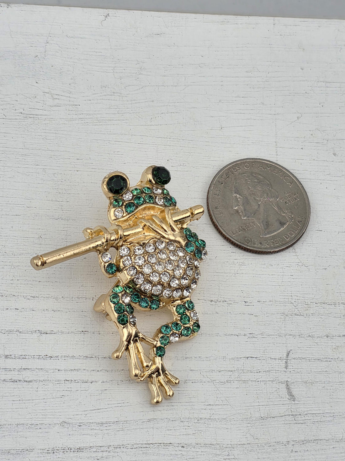 A decorative gold and green frog brooch with sparkling gemstones is displayed in a white ornate frame.