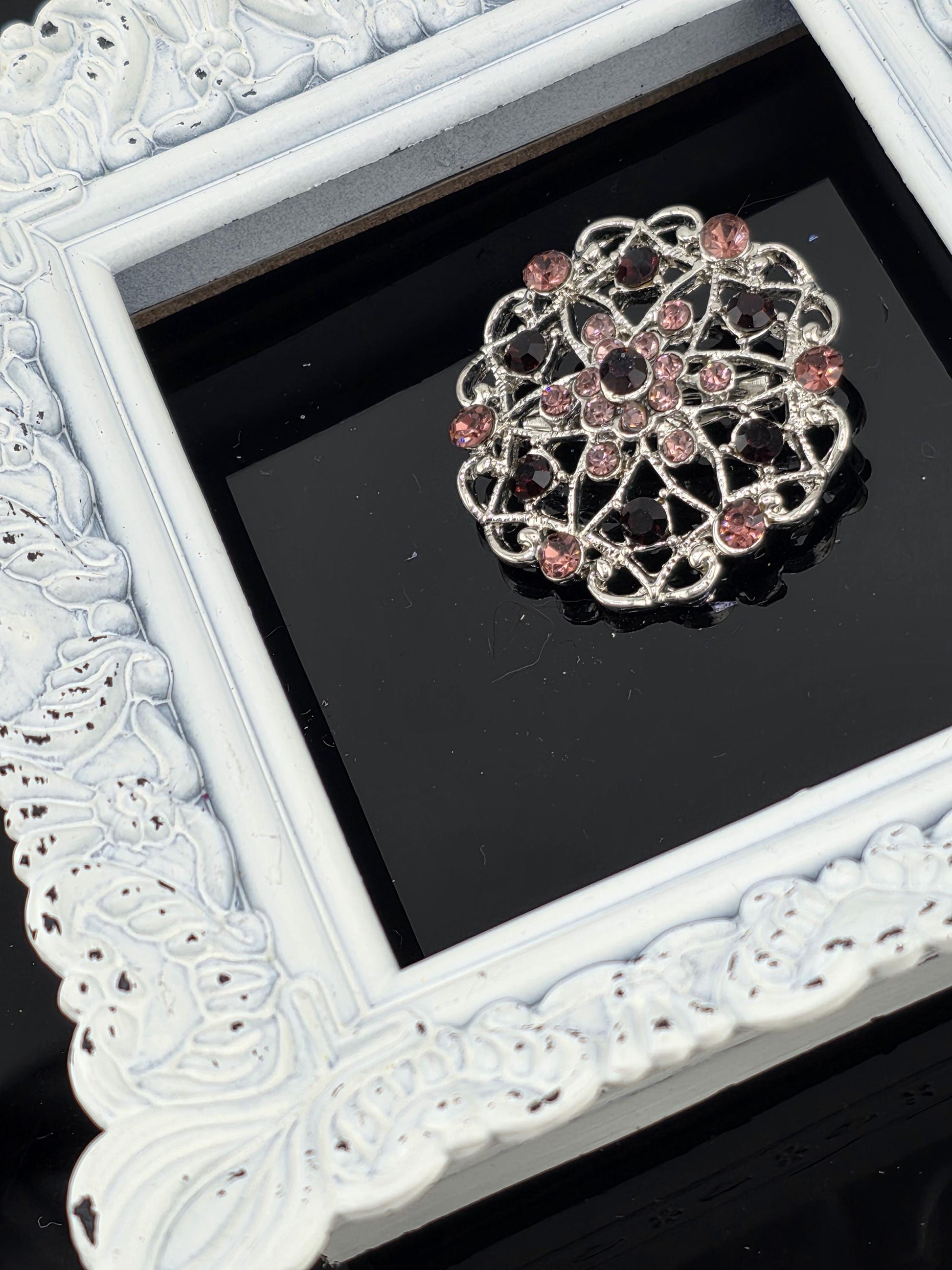 A decorative brooch with a floral design and pink gemstones is displayed in a white ornate frame against a black background.