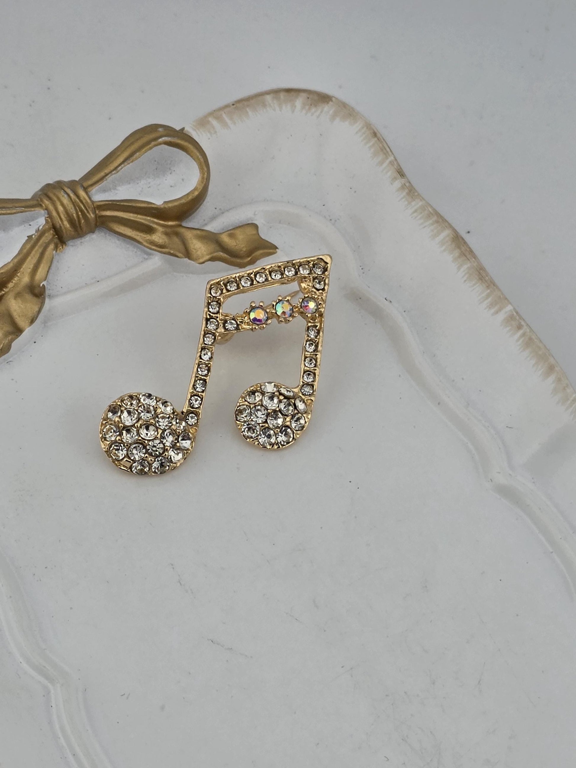 A decorative piece featuring a gold-colored ribbon and a large, ornate brooch with a musical note design, both adorned with sparkling rhinestones.
