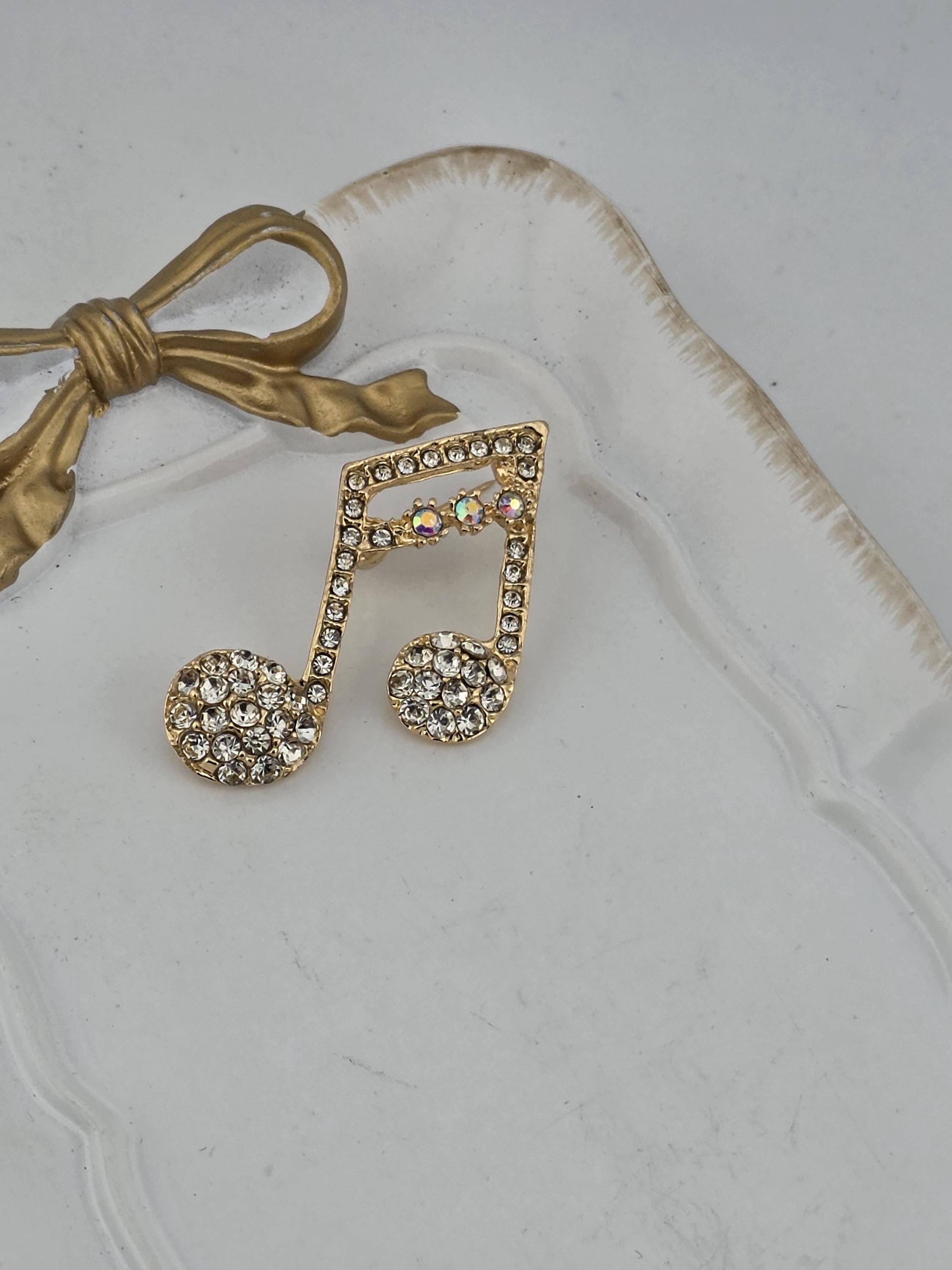 A decorative piece featuring a gold-colored ribbon and a large, ornate brooch with a musical note design, both adorned with sparkling rhinestones.