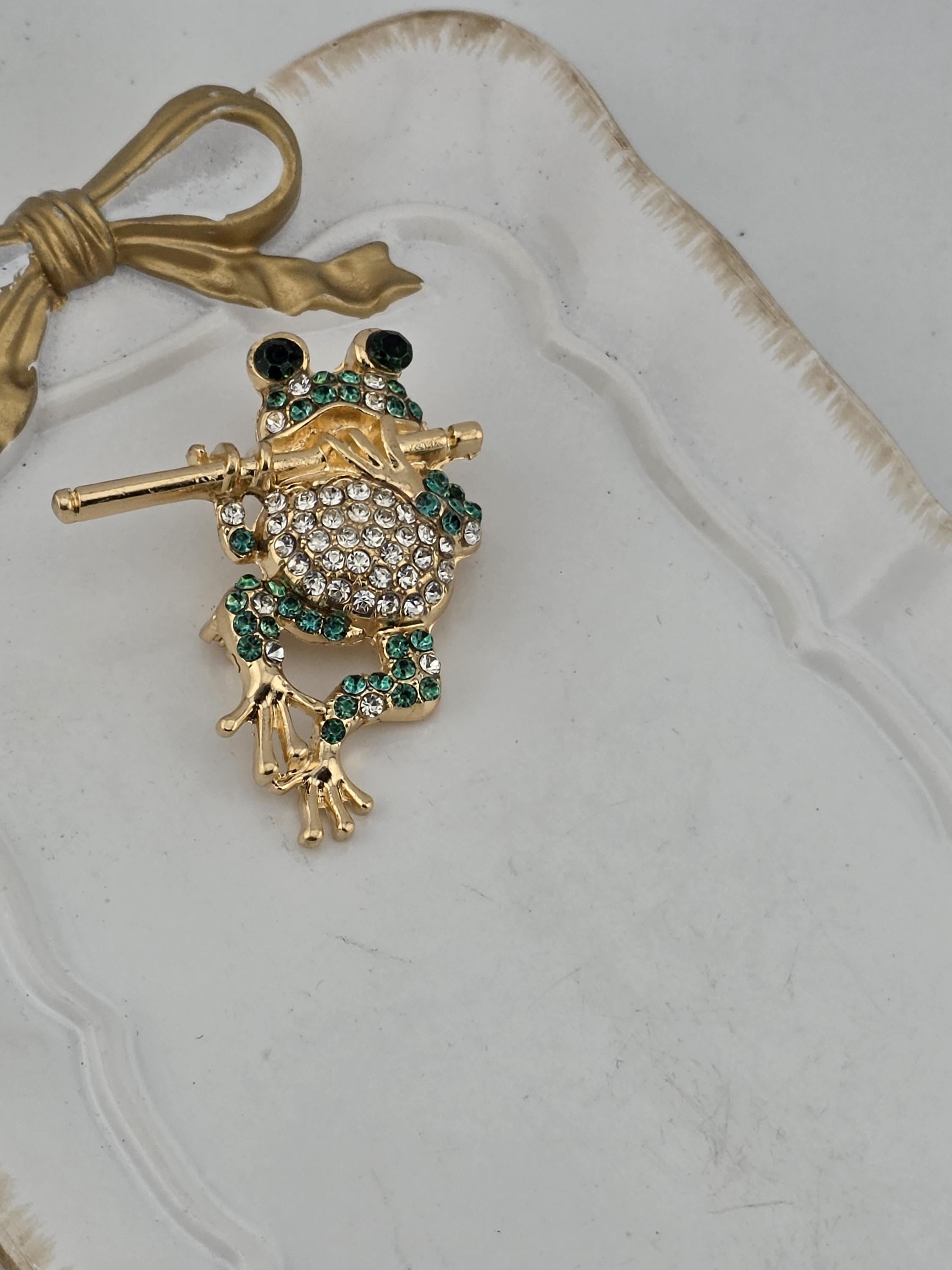 A decorative frog brooch with green gemstones and a gold-colored design, placed on a white surface.