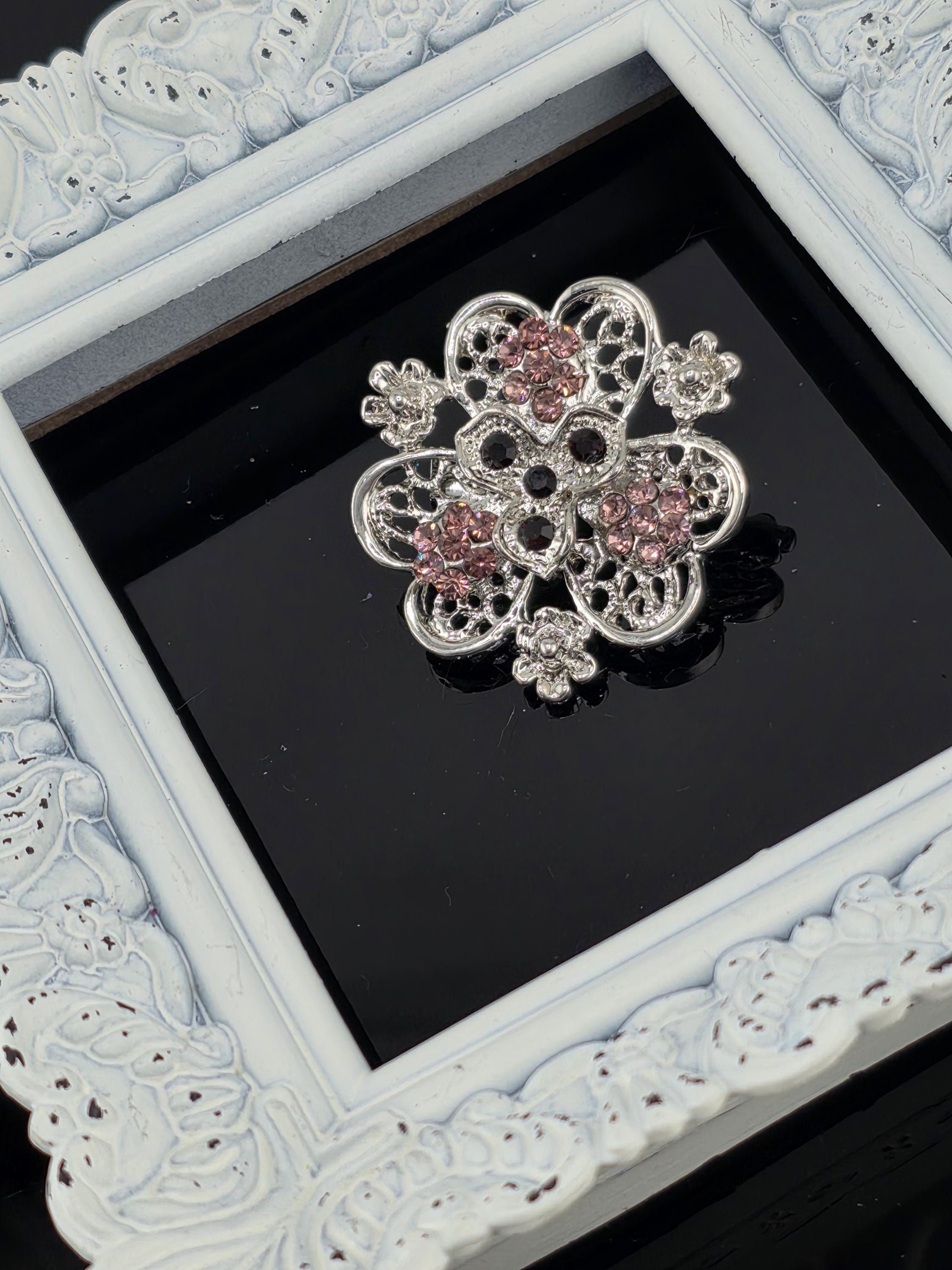 A decorative silver brooch with pink gemstones is displayed in a white ornate frame against a black background.