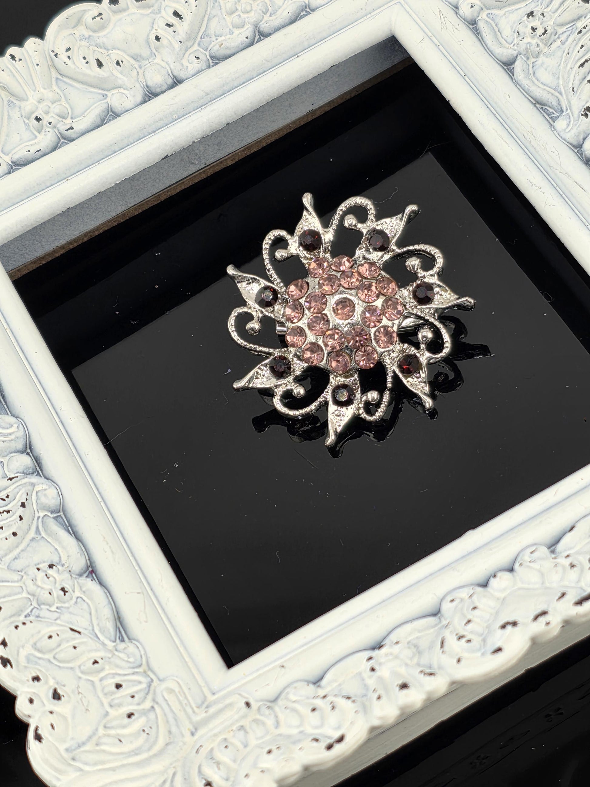 A decorative brooch with a floral design and sparkling gemstones is displayed within a white ornate frame against a black background.