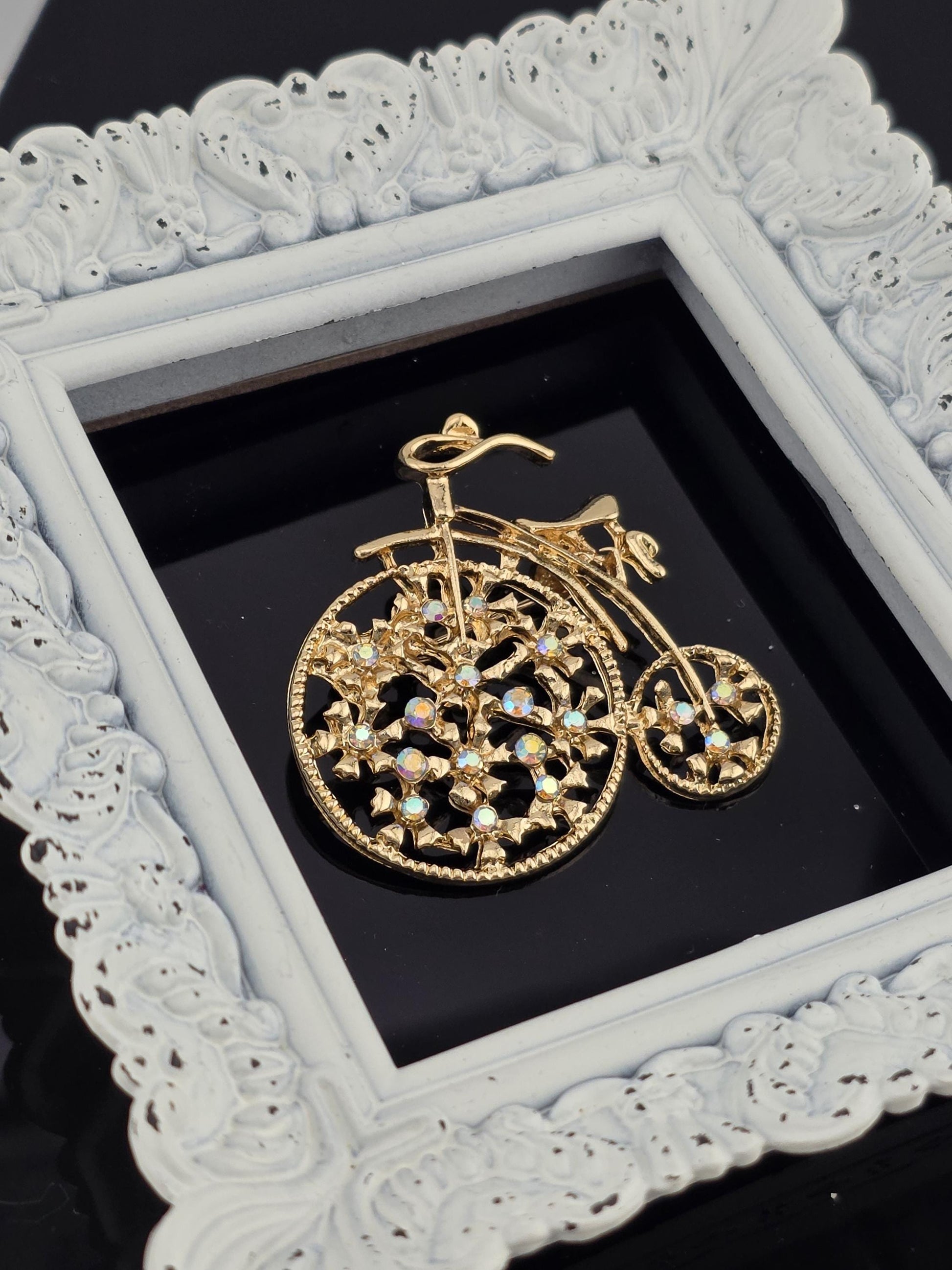 A decorative gold-colored bicycle brooch is displayed in a white ornate frame against a black background.