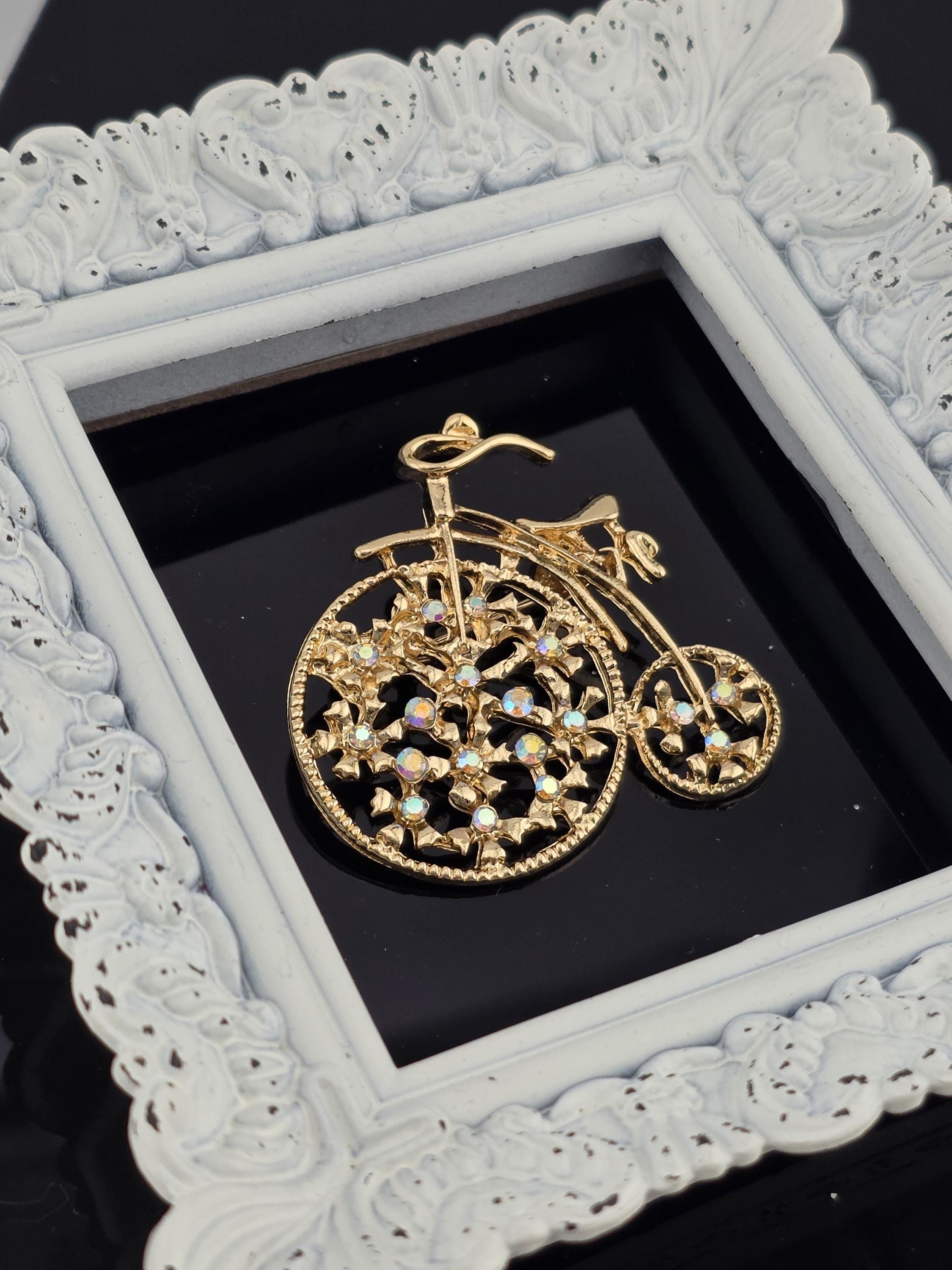 A decorative gold-colored bicycle brooch is displayed in a white ornate frame against a black background.