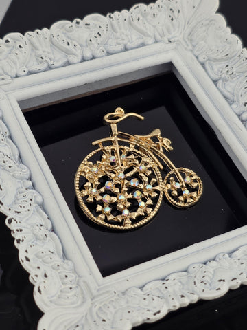 A decorative gold-colored bicycle brooch is displayed in a white ornate frame against a black background.
