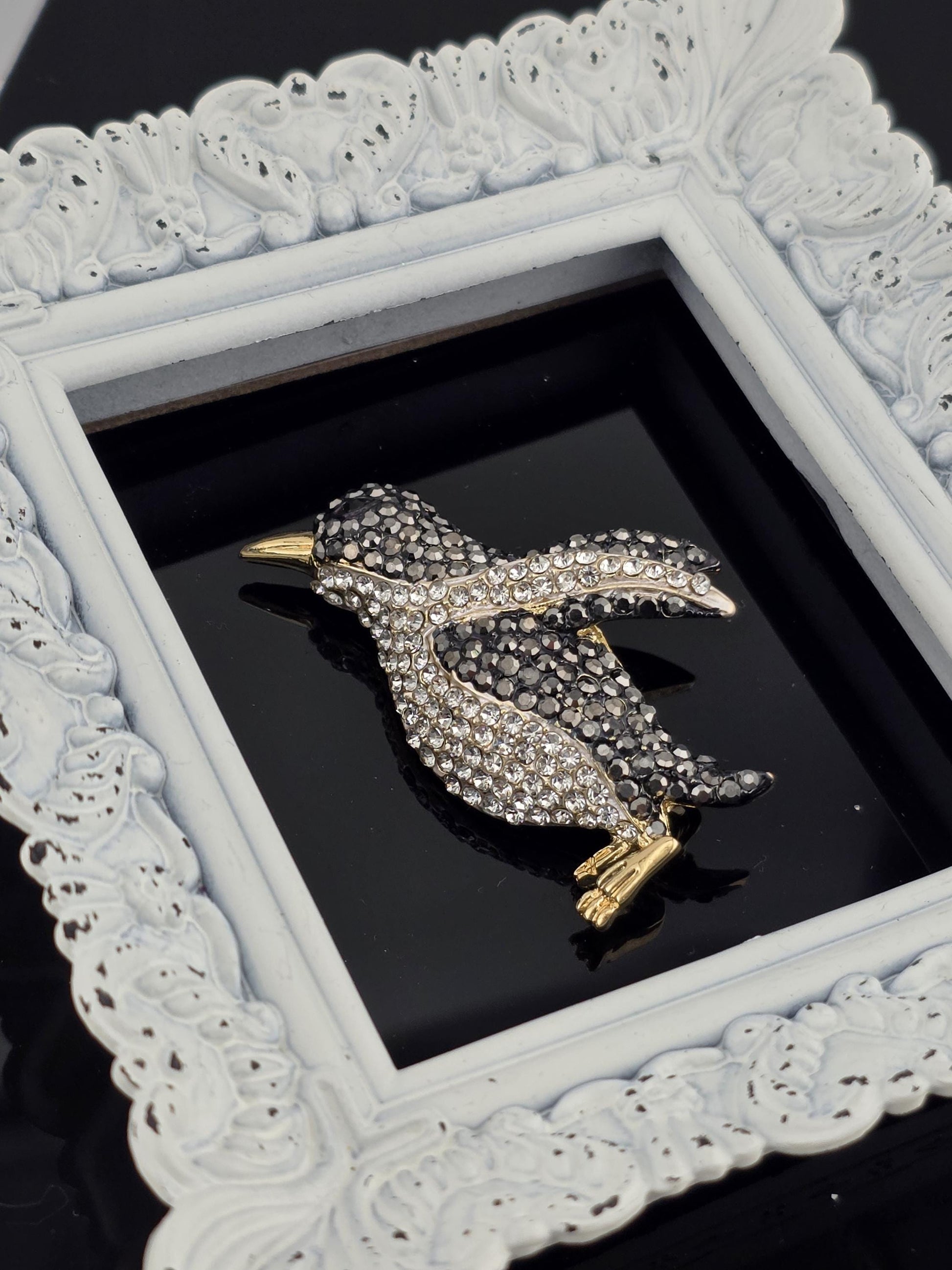 A decorative white frame with a black background, containing a sparkling rhinestone brooch in the shape of a bird.