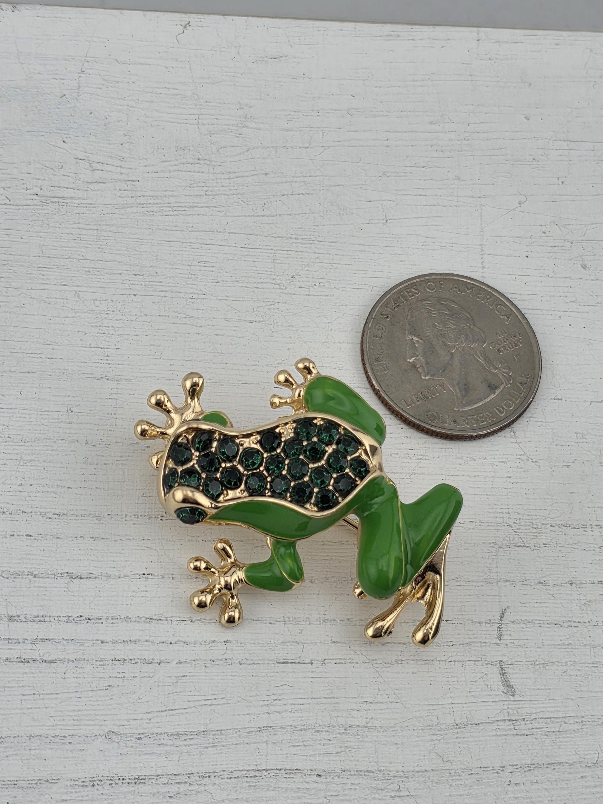 A green frog brooch with green gemstones is placed next to a US quarter on a white surface.