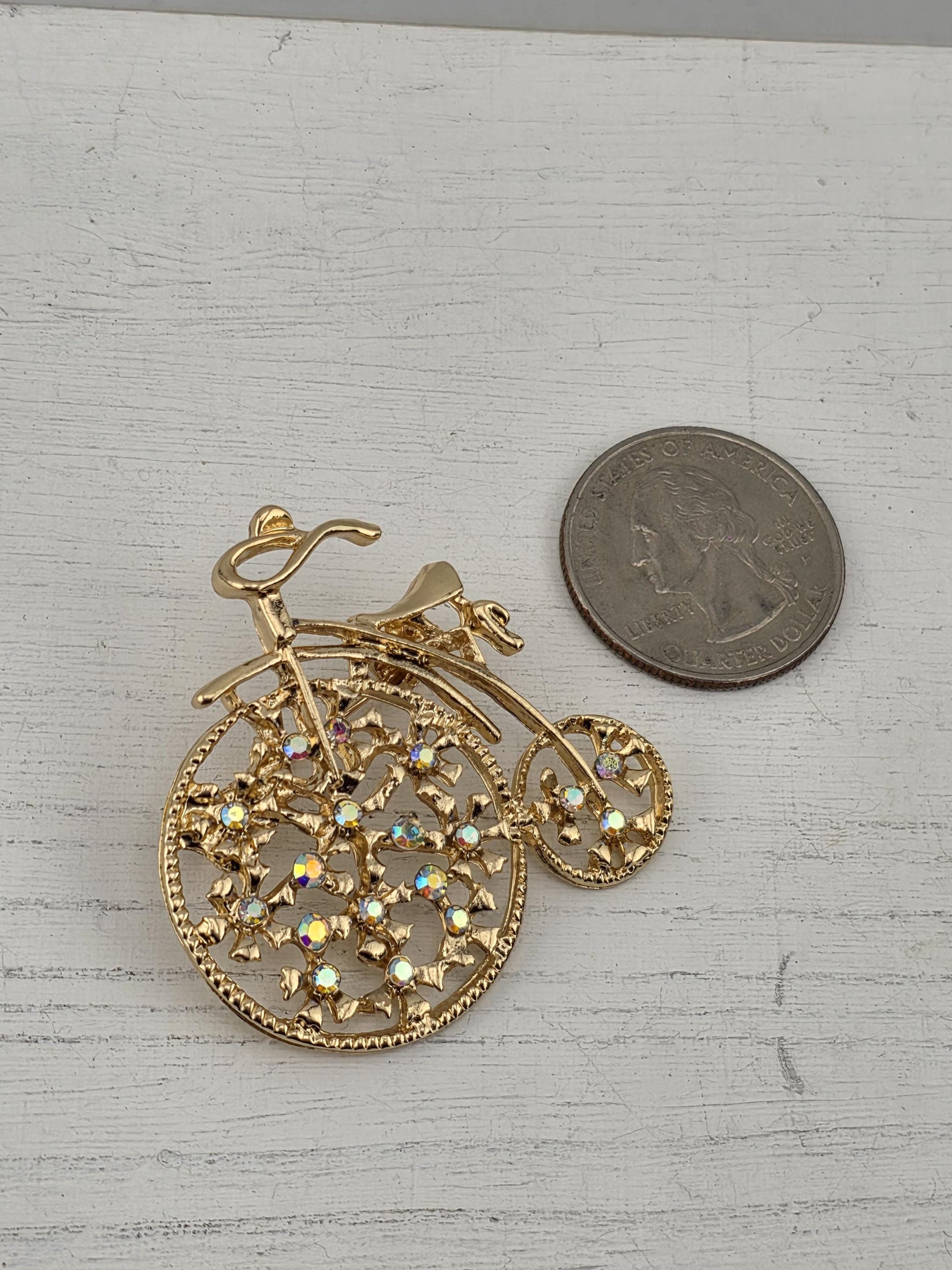A gold-colored brooch with a floral design and a small coin placed next to it on a white surface.