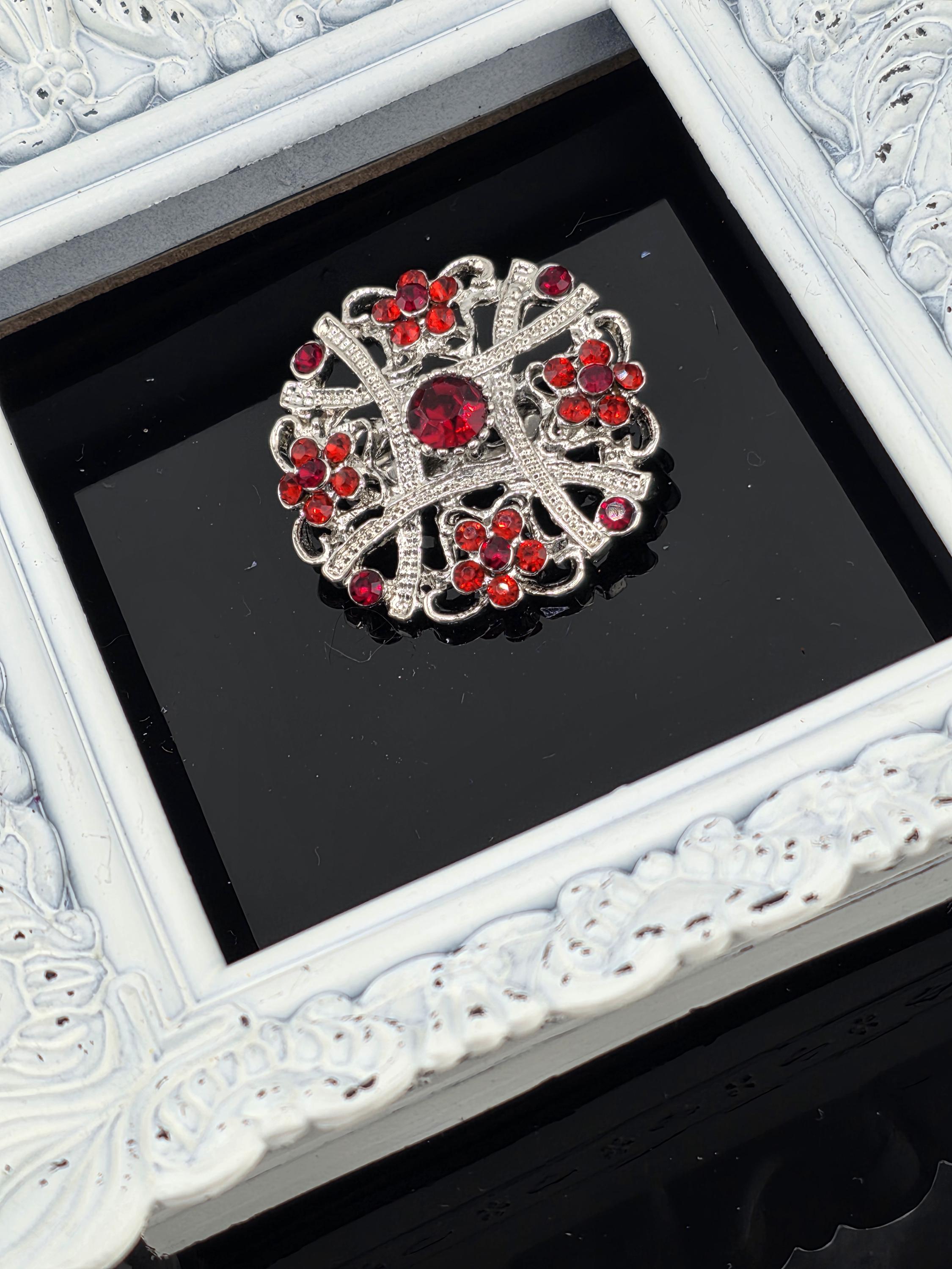 A decorative brooch with red gemstones is displayed in a white ornate frame against a black background.