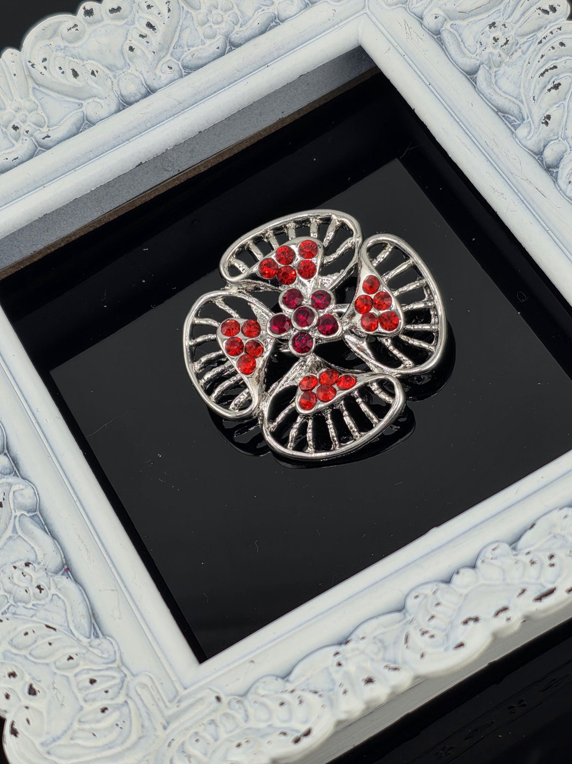 A decorative silver brooch with red gemstones is displayed in a white ornate frame against a black background.