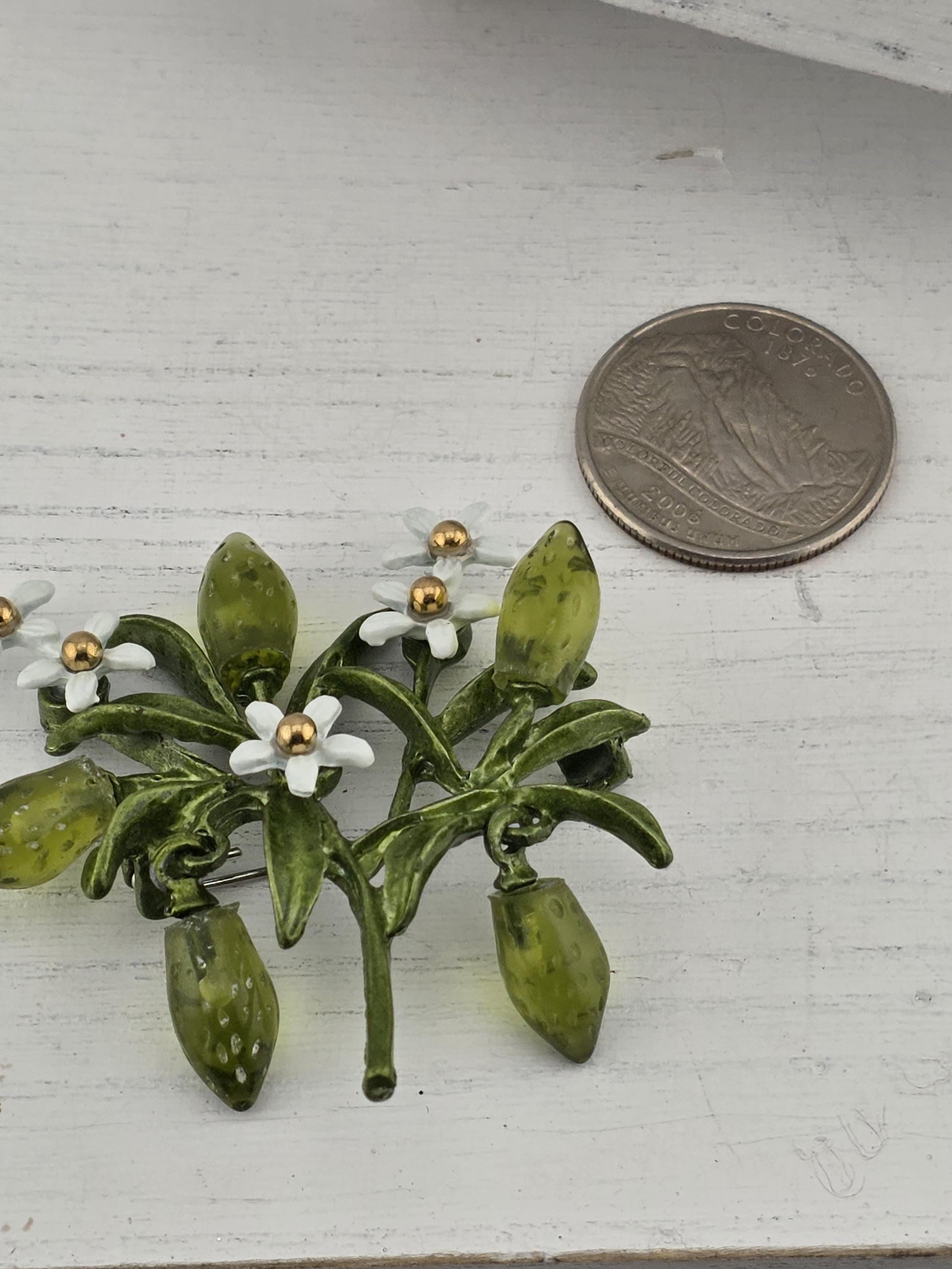 A close-up image of a green leaf-shaped brooch with white flowers, placed next to a silver coin on a white surface.