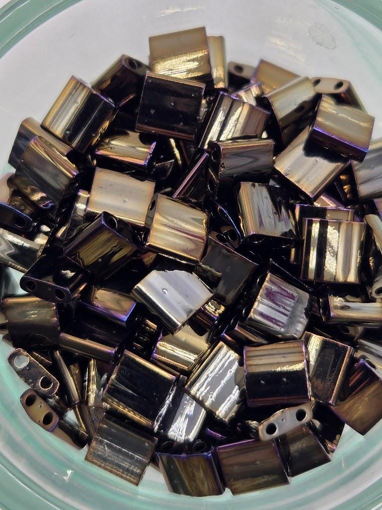 a close-up view of a collection of small, square-shaped metal pieces with a metallic sheen, possibly made of brass or bronze. The pieces are arranged in a pile, creating a visually interesting and textured composition.