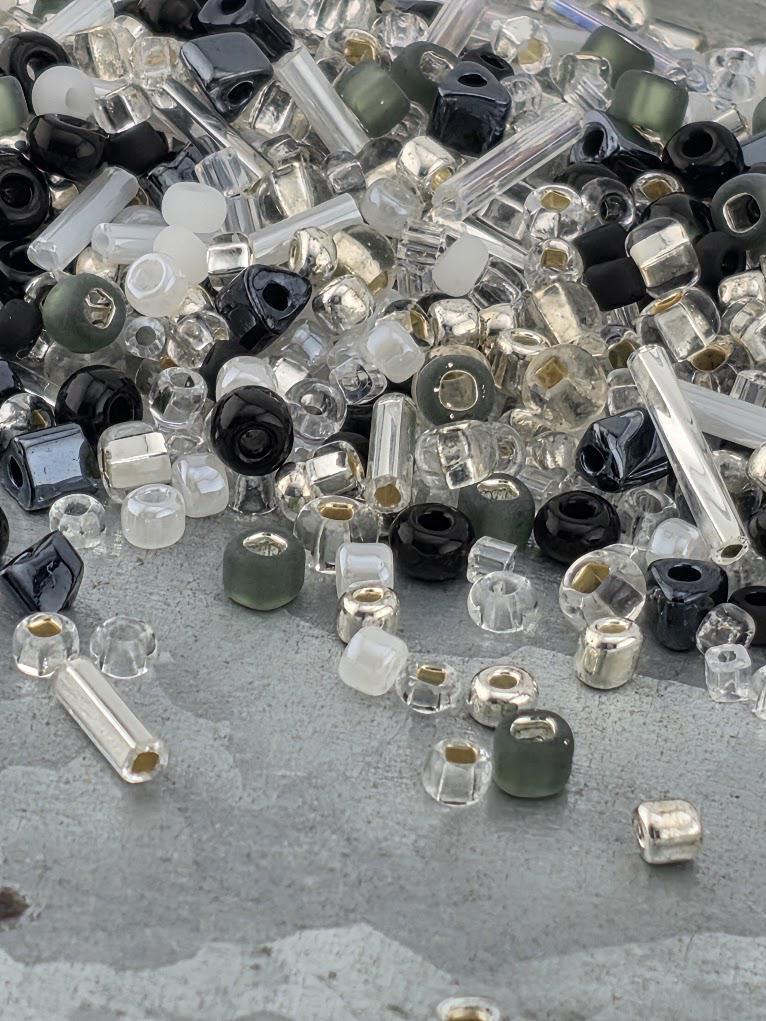 The image is a collection of various glass beads and glass rods scattered on a surface, with a mix of clear, black, and silver colors.