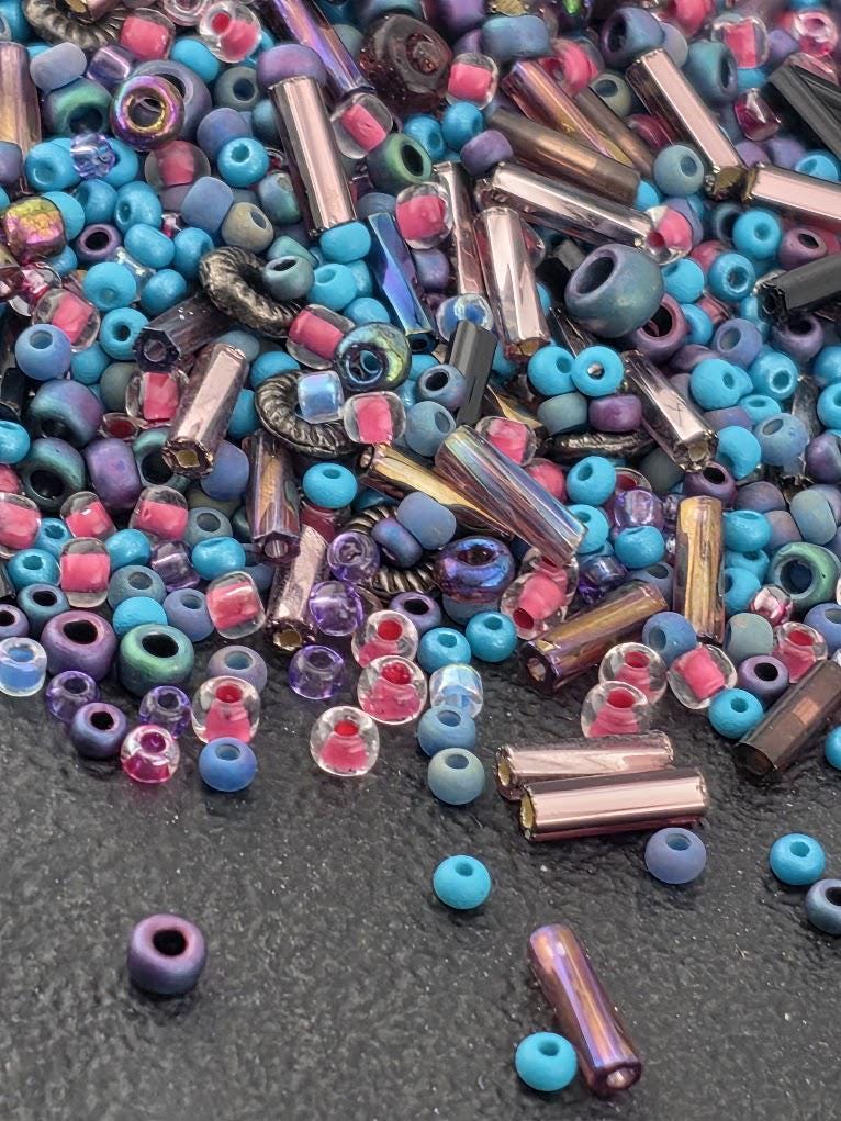 A colorful assortment of glass beads in various shapes and sizes, including clear, blue, purple, and multicolored glass beads, scattered on a dark surface.