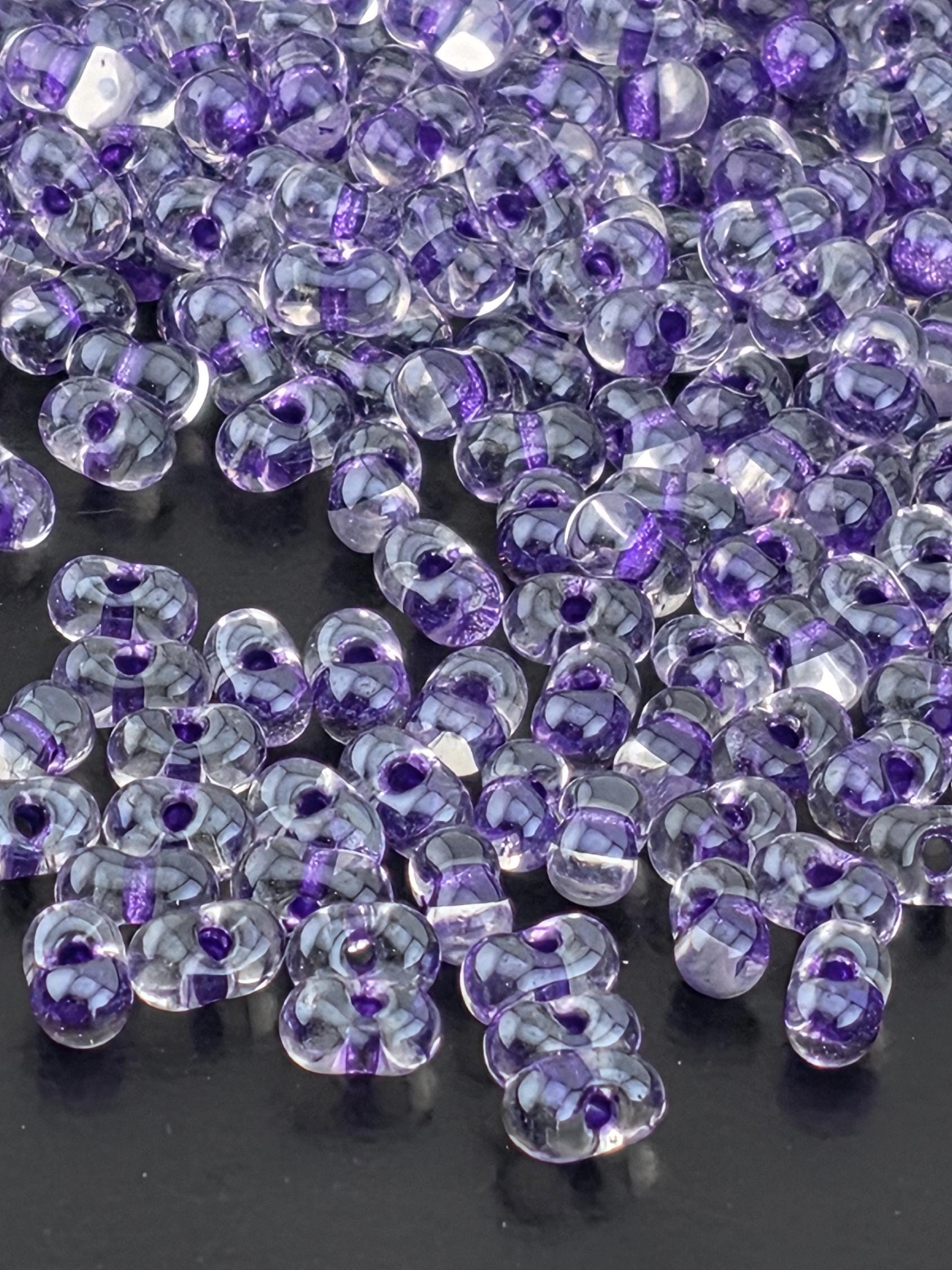 a close-up view of numerous clear glass beads scattered on a dark surface. The beads have a purple and white color scheme, creating a visually striking contrast against the dark background.