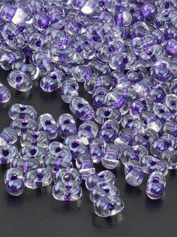 a close-up view of numerous clear glass beads scattered on a dark surface. The beads have a purple and white color scheme, creating a visually striking contrast against the dark background.