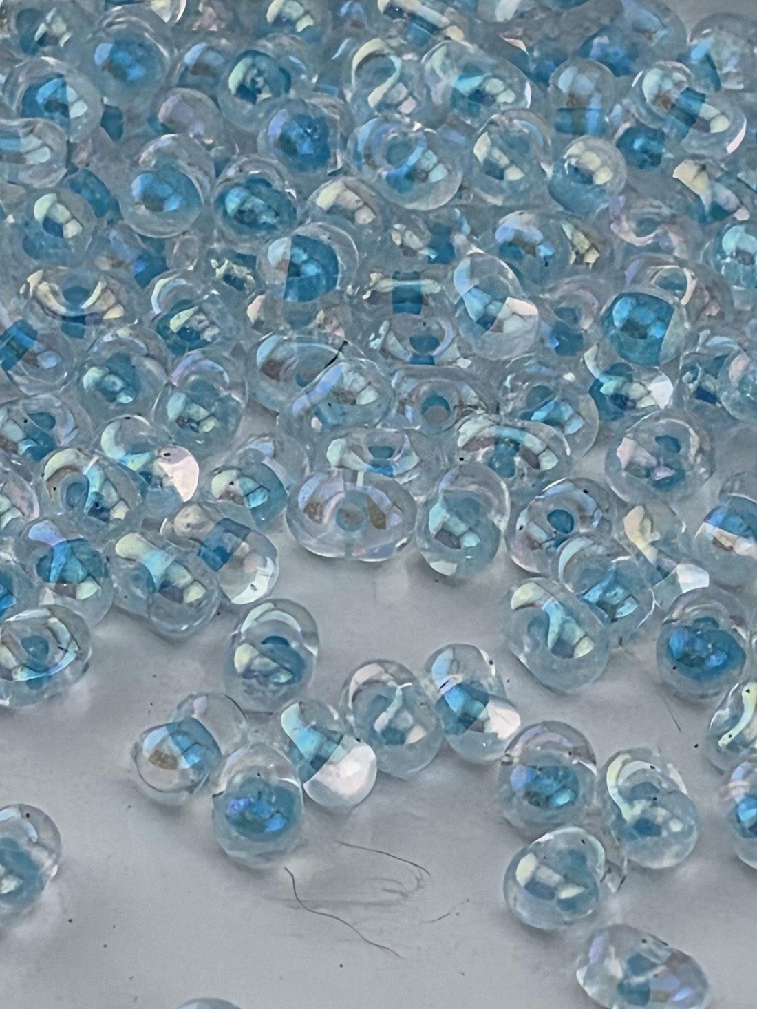 a close-up view of numerous clear, transparent beads scattered on a surface. The beads appear to be made of glass and have a blue-green tint, giving them a unique and iridescent appearance. The image does not contain any text or specific details about the beads, and it does not depict any actions or movements. The focus is on the individual beads and their arrangement on the surface.