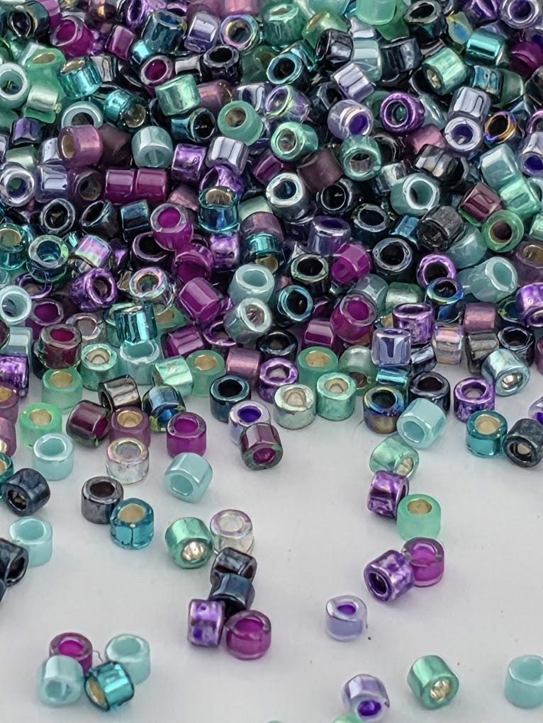 A colorful pile of multicolored beads, including shades of purple, blue, green, and silver, scattered across a white surface.