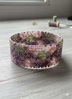 A beaded bracelet with a colorful, abstract pattern of pink, green, and purple beads is displayed on a wooden surface.