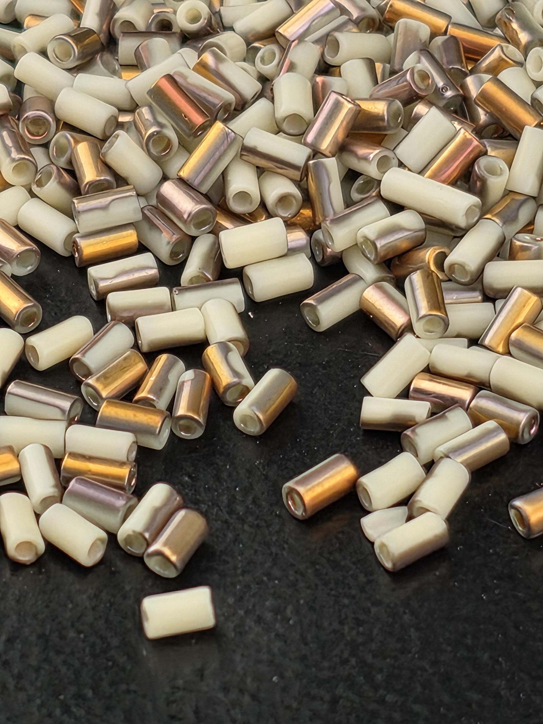 a close-up view of a pile of small, cylindrical objects with a mix of white and gold colors. The objects appear to be made of a shiny material, possibly metal or glass, and are scattered across a dark surface.